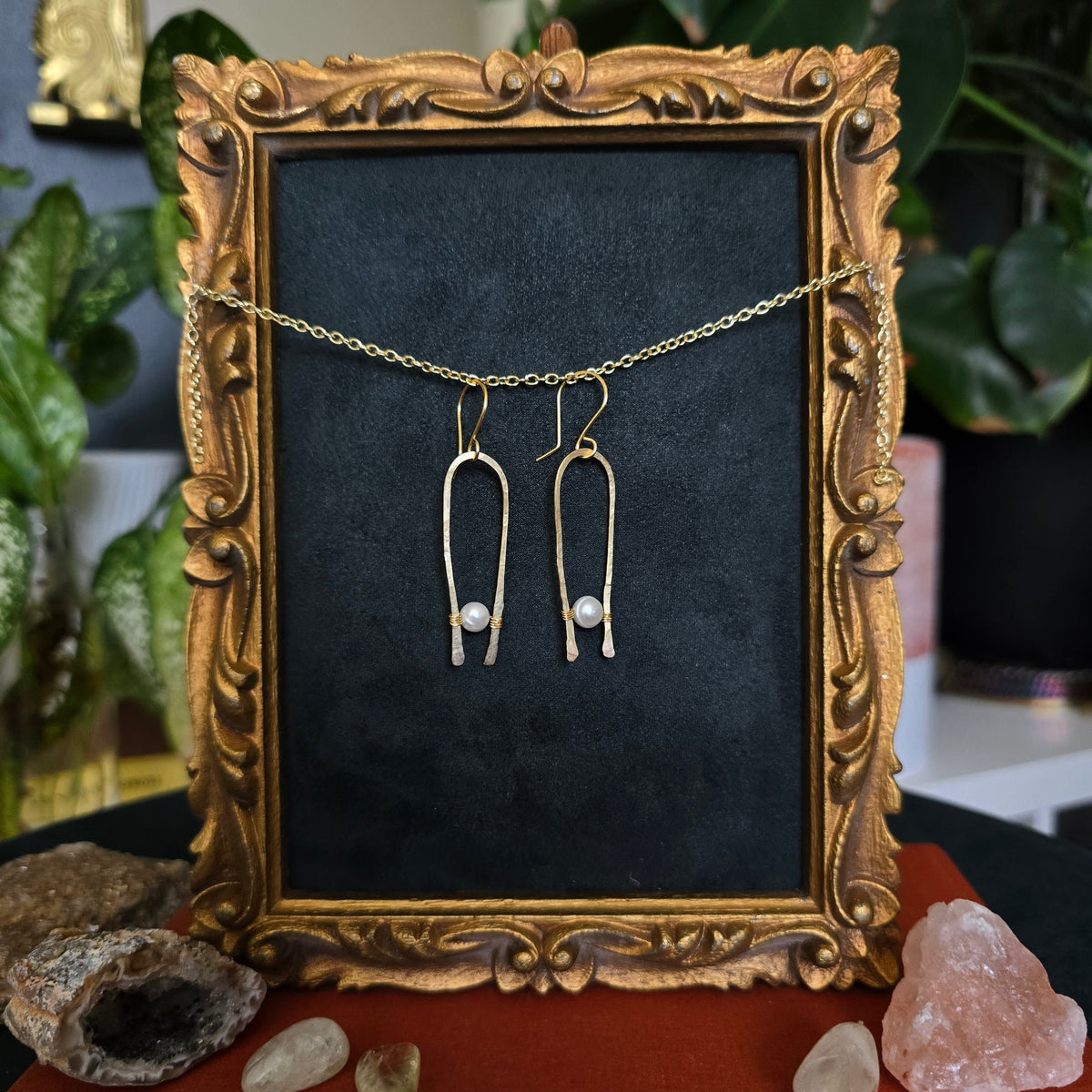 Gold-toned dangling earrings with thin chain drops and small pearl-like beads, displayed on a dark velvet insert inside an ornate gold picture frame.