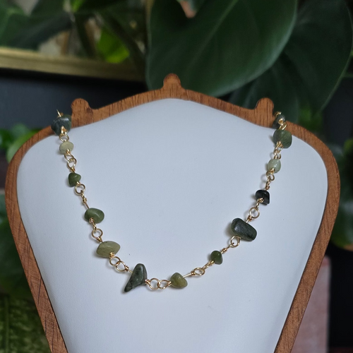A beaded necklace with olive green and dark brown stone beads on a gold chain, displayed on a white jewelry bust with a wooden frame. The bust is set on a wooden stand with a blurred background of green houseplants.