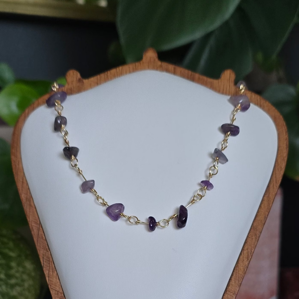 A beaded necklace with purple, white, and gold beads displayed on a white jewelry bust with a wooden frame, set on a wooden stand in a styled indoor setting with plants and crystals.