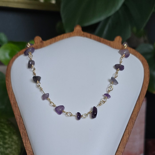 A beaded necklace with purple, white, and gold beads displayed on a white jewelry bust with a wooden frame, set on a wooden stand in a styled indoor setting with plants and crystals.