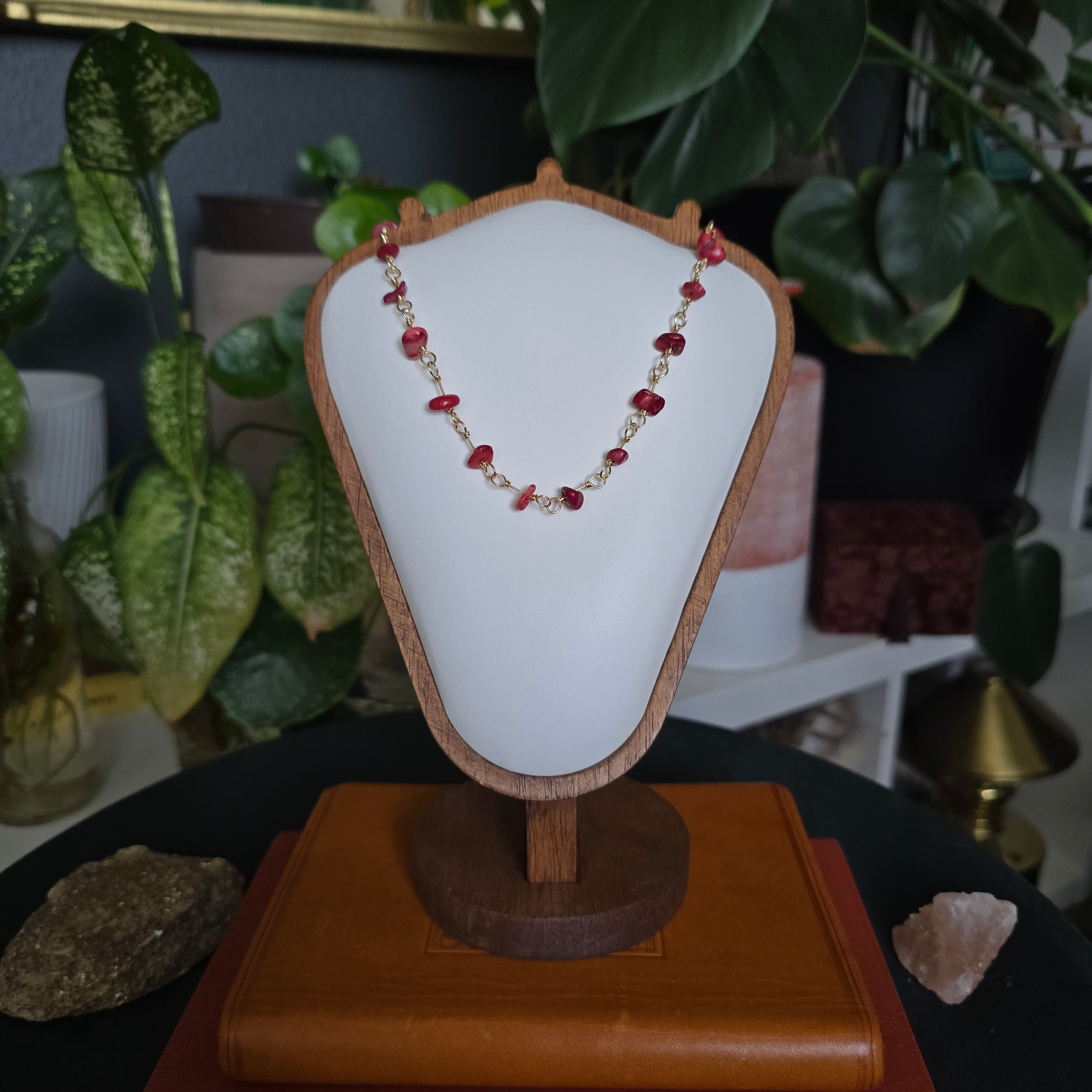 A beaded necklace with red and gold beads displayed on a white jewelry bust stand, set against a background of green houseplants and decorative crystals.