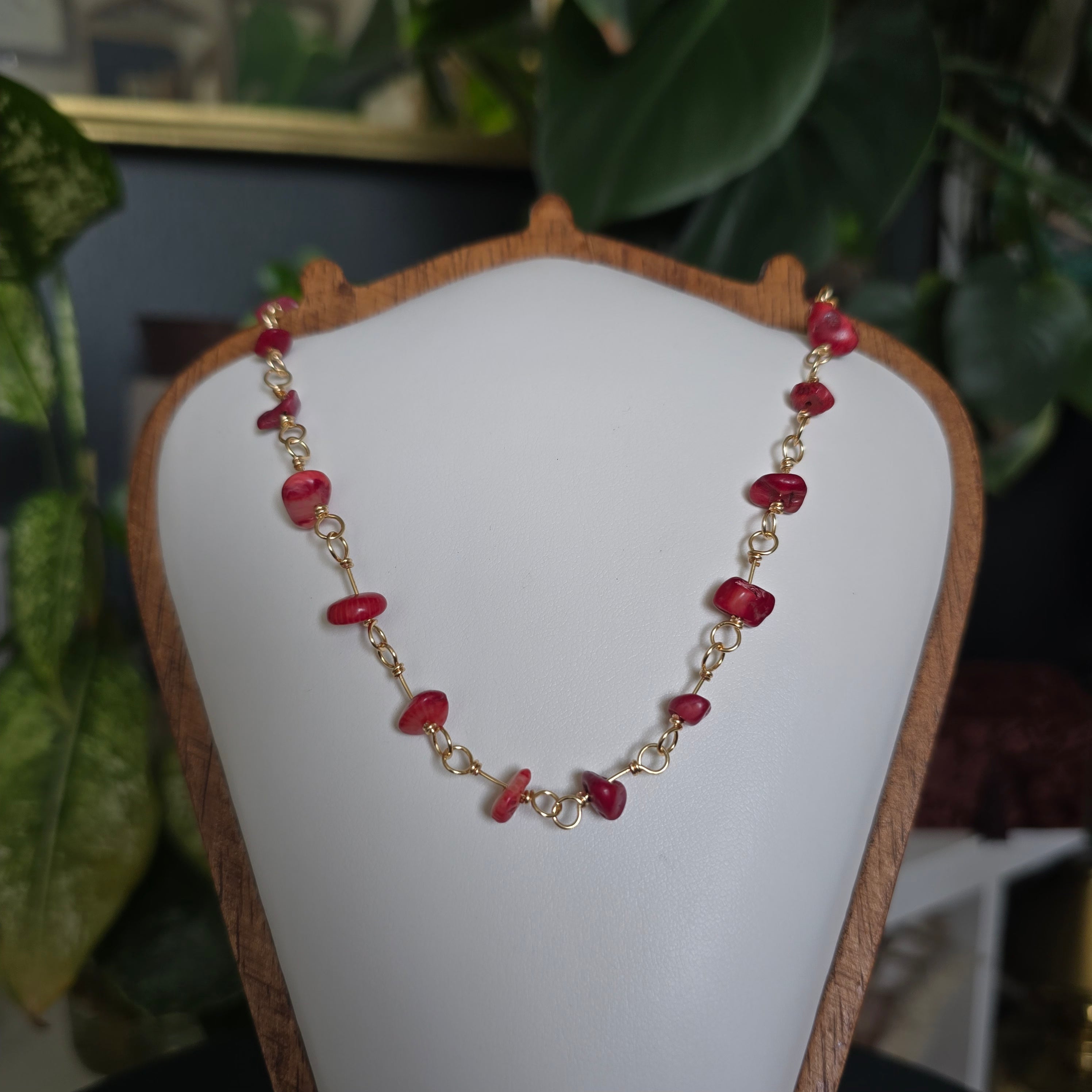 A gold chain necklace with small red faceted gemstone beads, displayed on a white jewelry bust with green plants in the background.