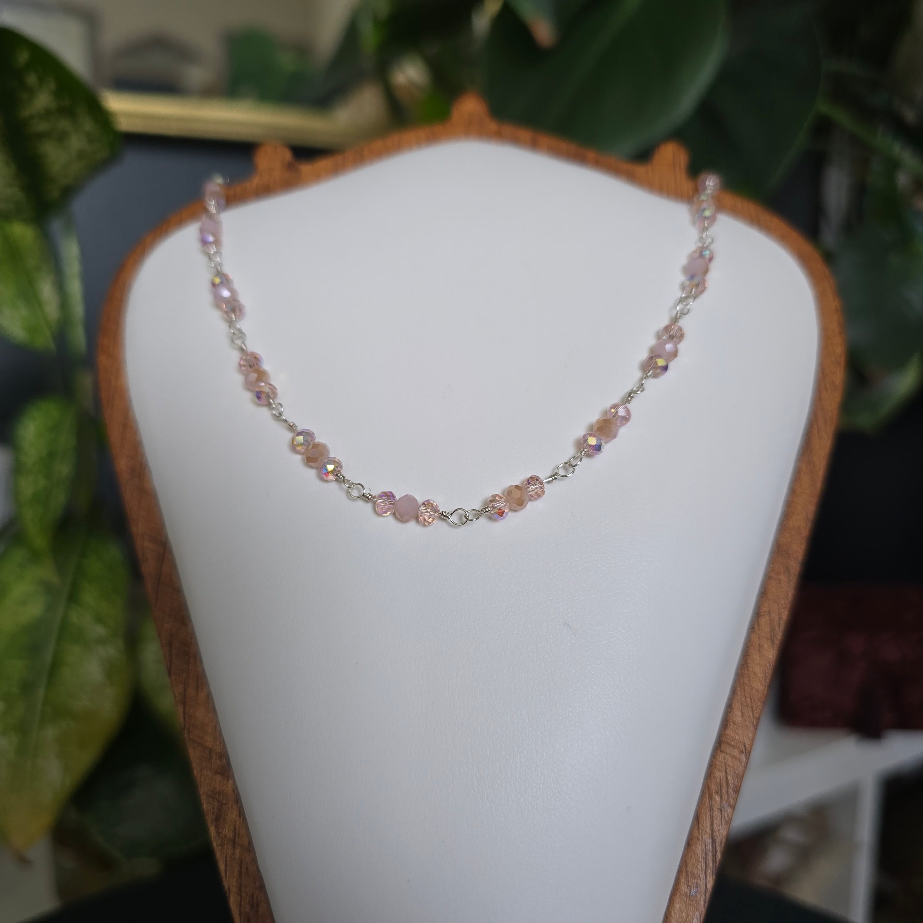 A delicate beaded necklace with small iridescent pink and peach beads separated by silver-toned links, displayed on a white jewelry bust with a wooden frame. Green houseplants are visible in the softly blurred background.