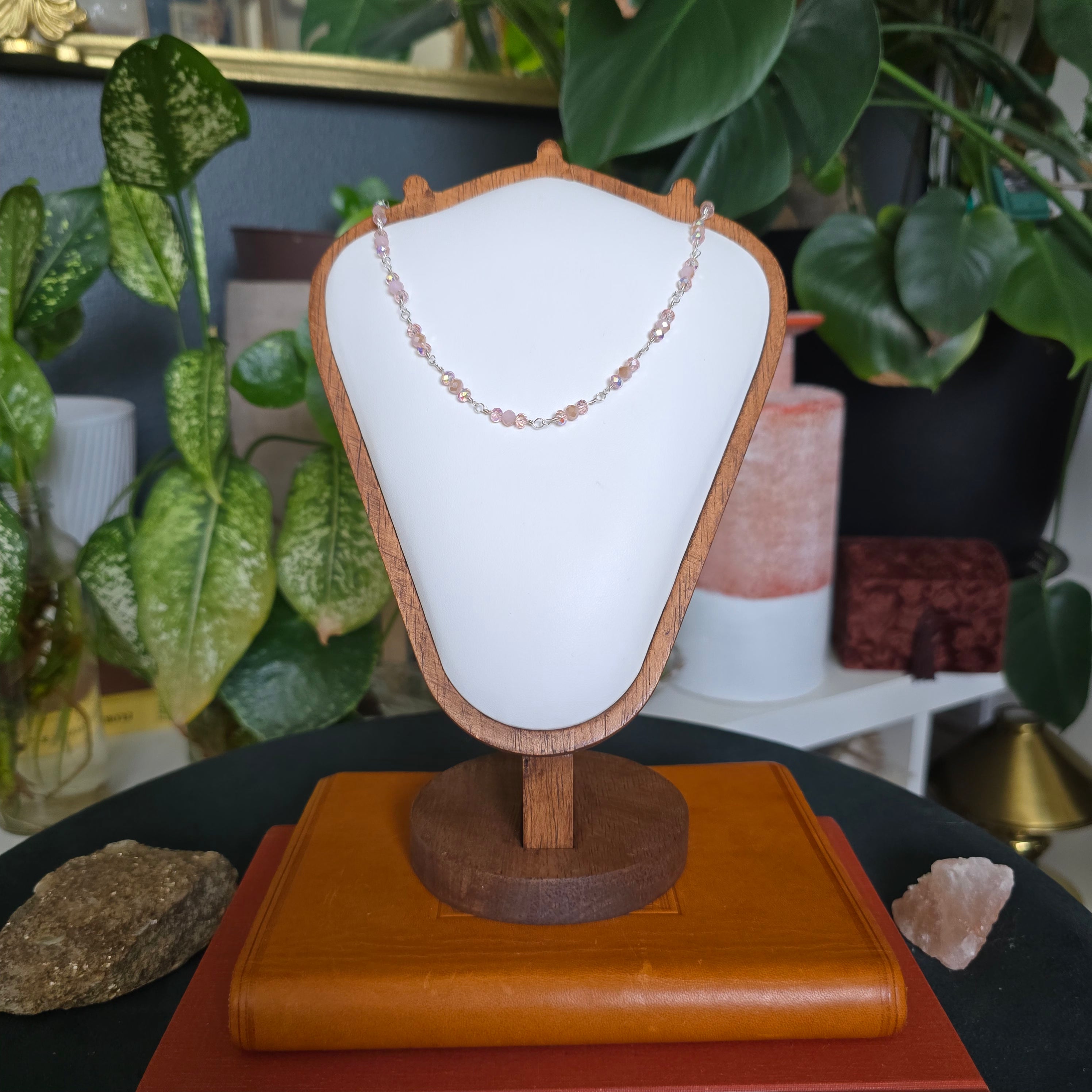 A delicate beaded necklace in soft pink tones displayed on a white jewelry bust with a wooden frame, set on a wooden stand. The bust is placed on a book on a dark table, surrounded by green houseplants and crystals.