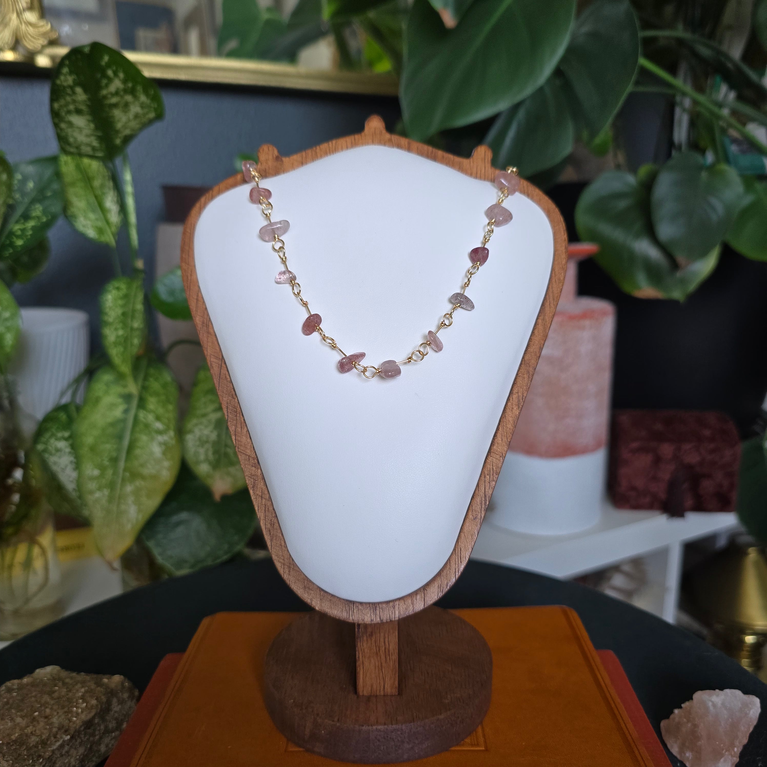A delicate gold chain necklace with small pink and reddish stone chips, displayed on a white bust stand with a wooden frame. The stand is placed on an orange book, with a dark table, plants, and decorative items in the background.
