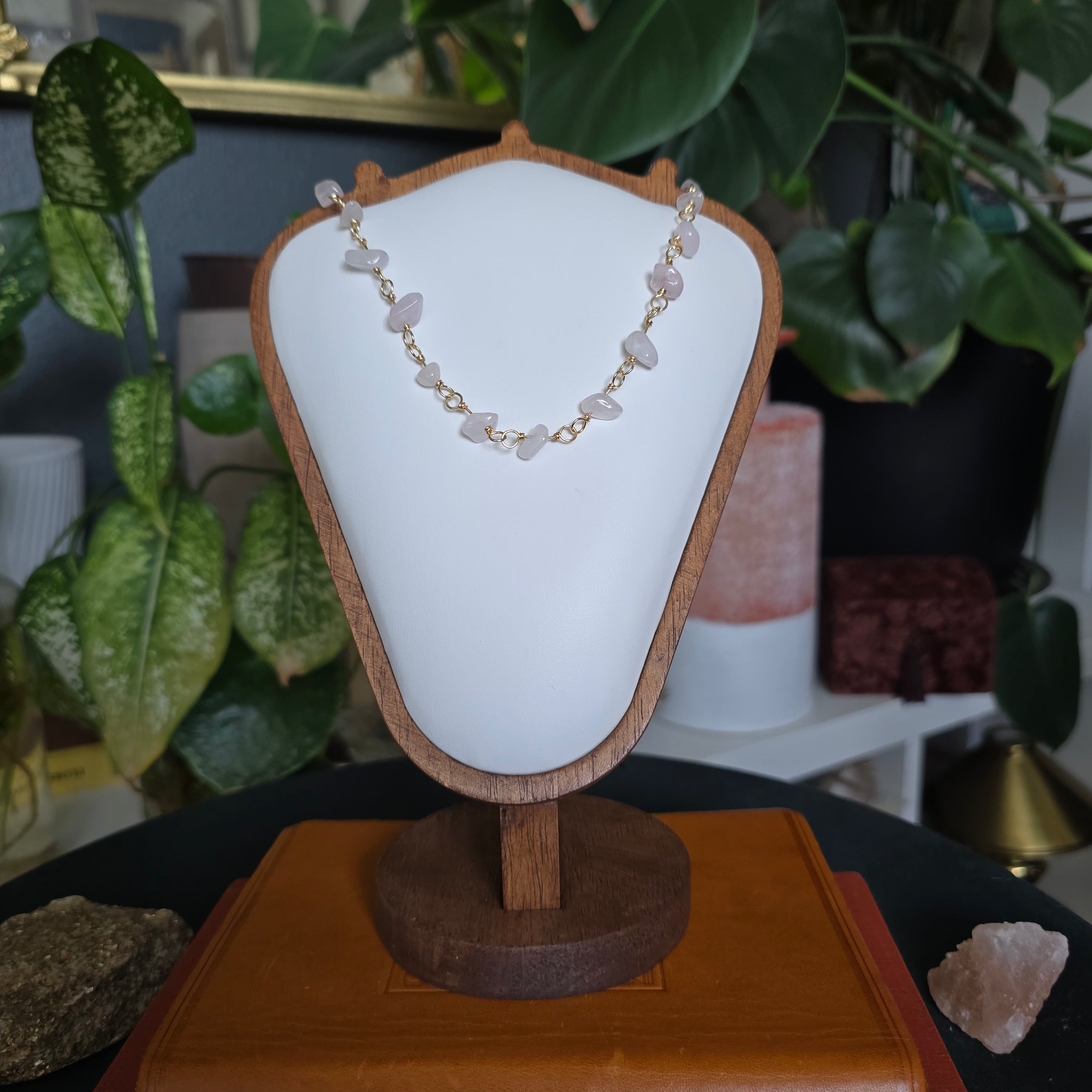A gold chain necklace with light pink stone chips displayed on a white cone jewelry bust with a wooden frame, set on a wooden stand in a styled indoor setting with plants and crystals.