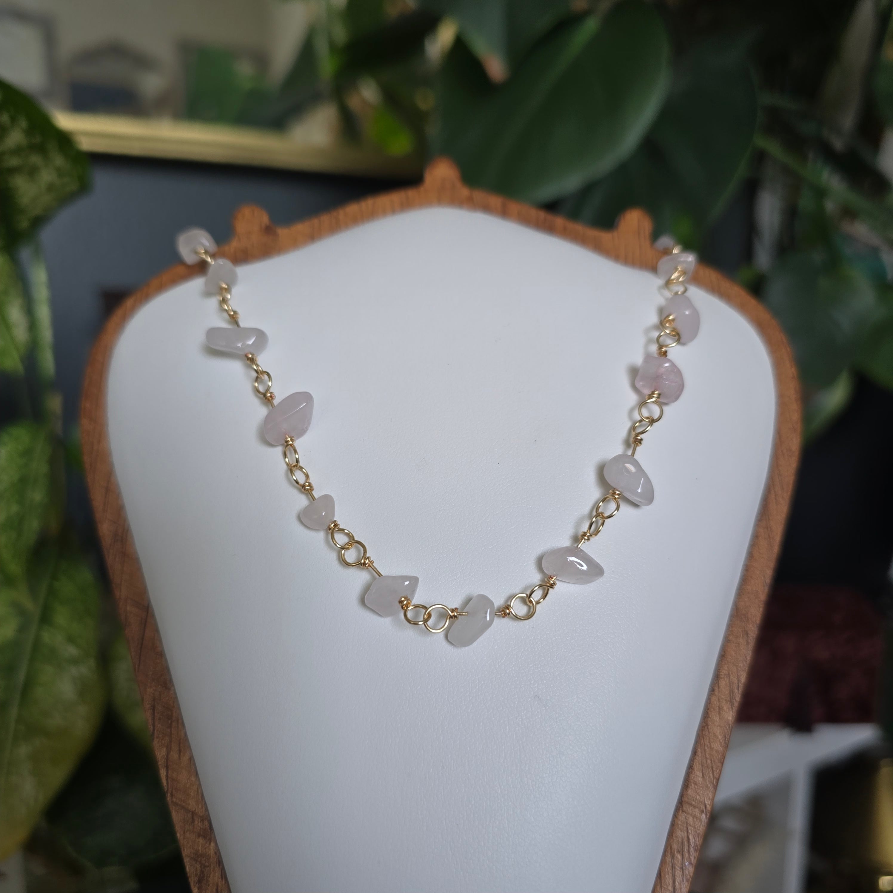 A gold chain necklace with irregular pale pink stone beads, displayed on a white jewelry bust with a wooden frame. The background features a blurred indoor setting with green plants.