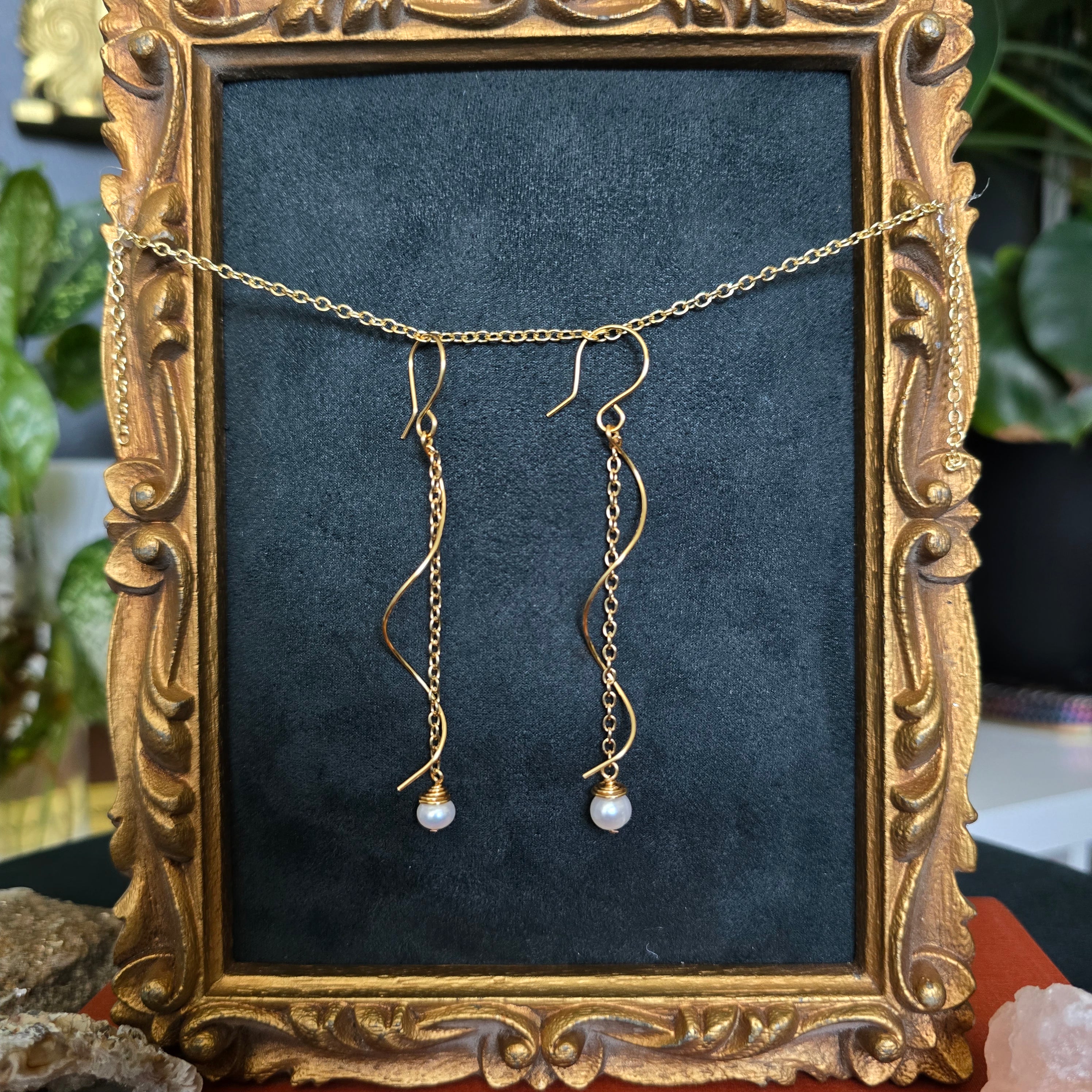 Gold-toned dangling earrings with thin chain drops and small pearl-like beads, displayed on a dark velvet insert inside an ornate gold picture frame.