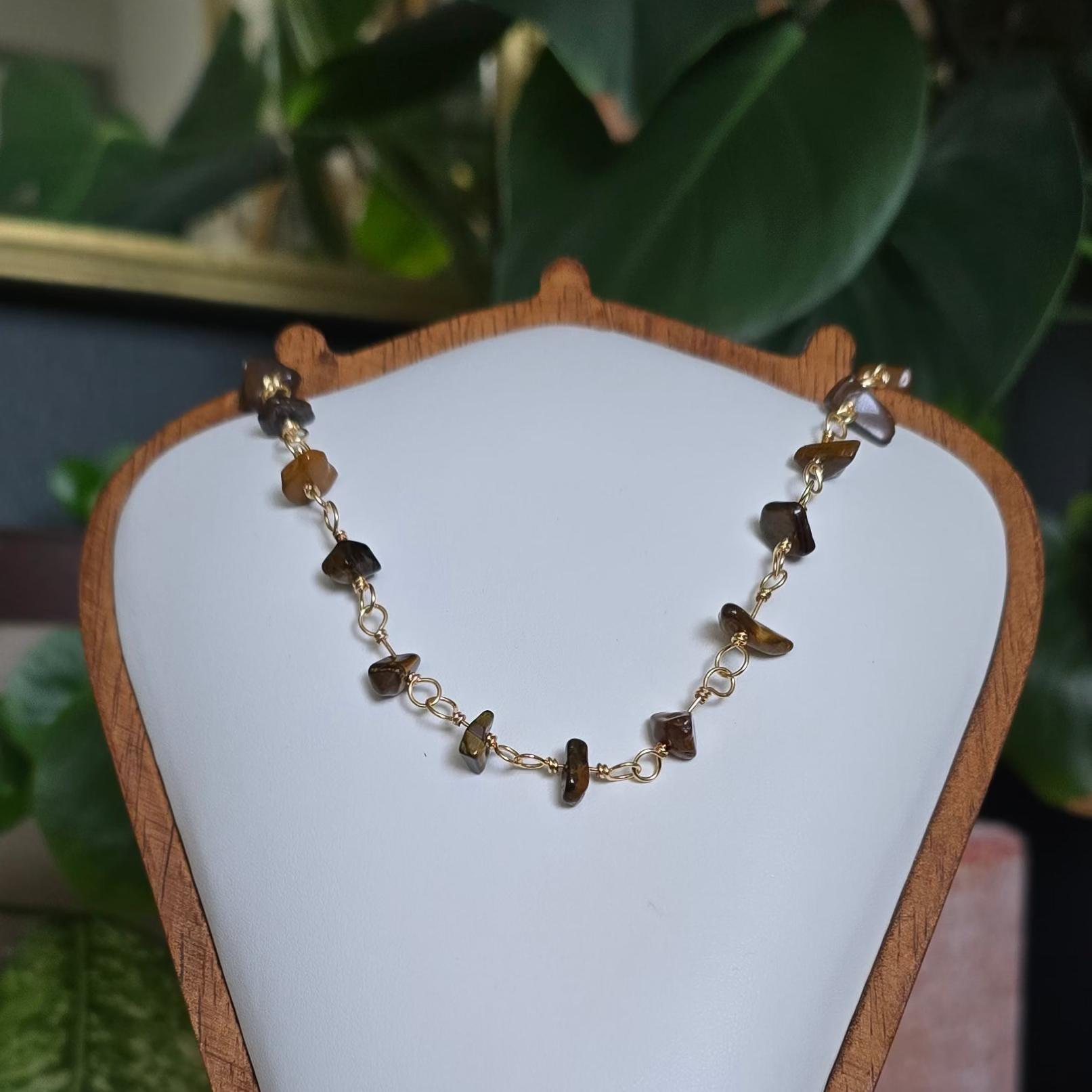 A gold chain necklace with small brown and amber stone beads displayed on a white jewelry bust stand, set against a background of green houseplants.