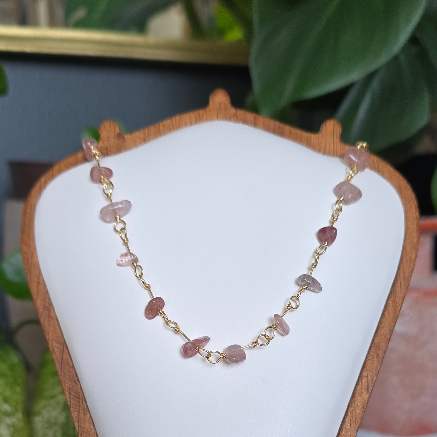 A delicate gold chain necklace with small pink and reddish stone beads displayed on a white jewelry bust stand, set against a styled background with green plants and crystals.