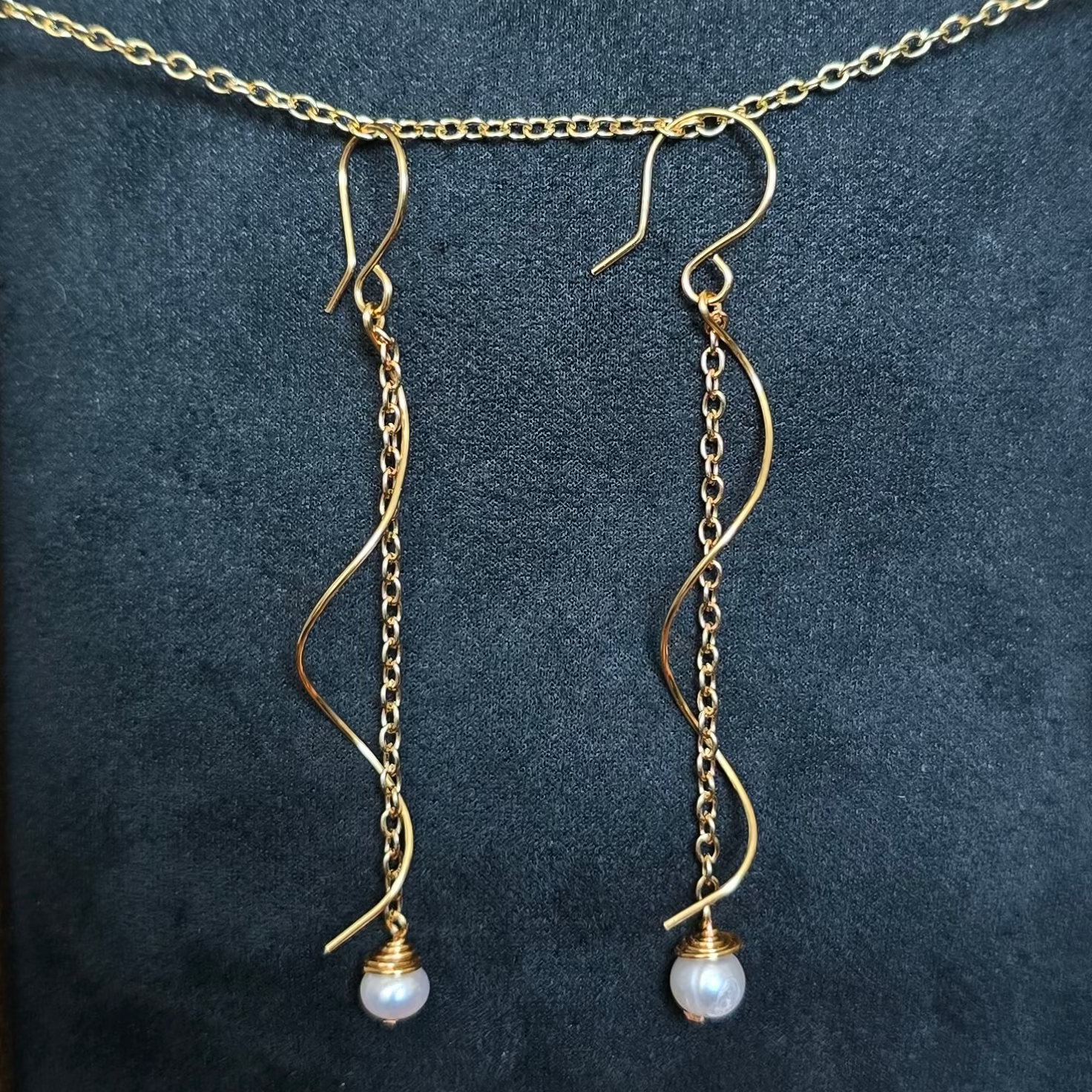 Gold dangle earrings with curved wire-like chains and small white pearl drops, displayed on a black velvet surface inside an ornate gold picture frame. The frame is set against a softly blurred indoor background with plants and decor.